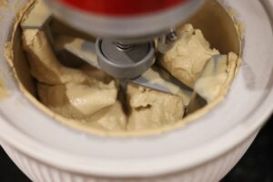 Coffee infused ice-cream base in a kitchen aid bowl attachment, with a red colour to the machine in a white bowl. The base is mid churn.