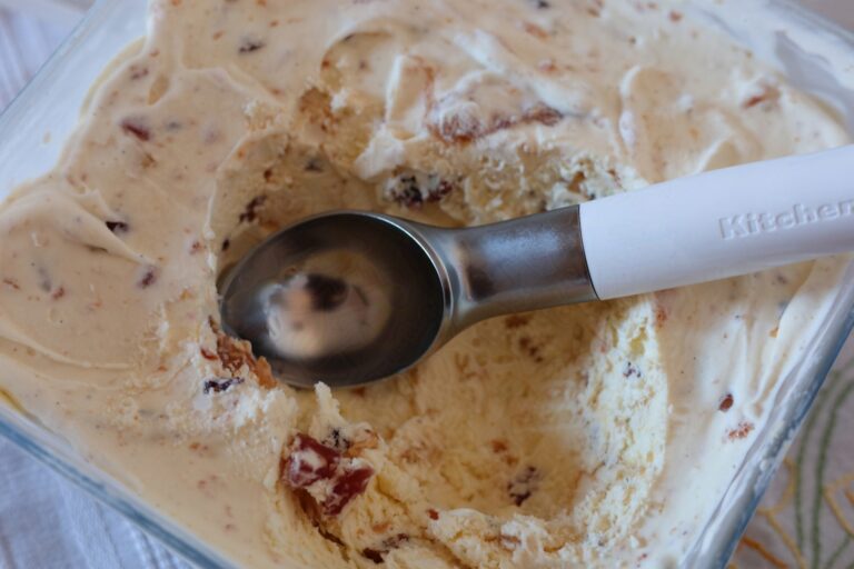 Classic Tutti Frutti ice cream. It is sat on a white cloth in a glass freezer dish. It has a scoop taken out of it, with a white and silver ice cream scoop sat in the scoops now gone. You can see some of the fruit, nut and paste throughout.