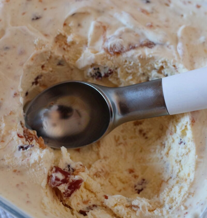 Classic Tutti Frutti ice cream. It is sat on a white cloth in a glass freezer dish. It has a scoop taken out of it, with a white and silver ice cream scoop sat in the scoops now gone. You can see some of the fruit, nut and paste throughout.